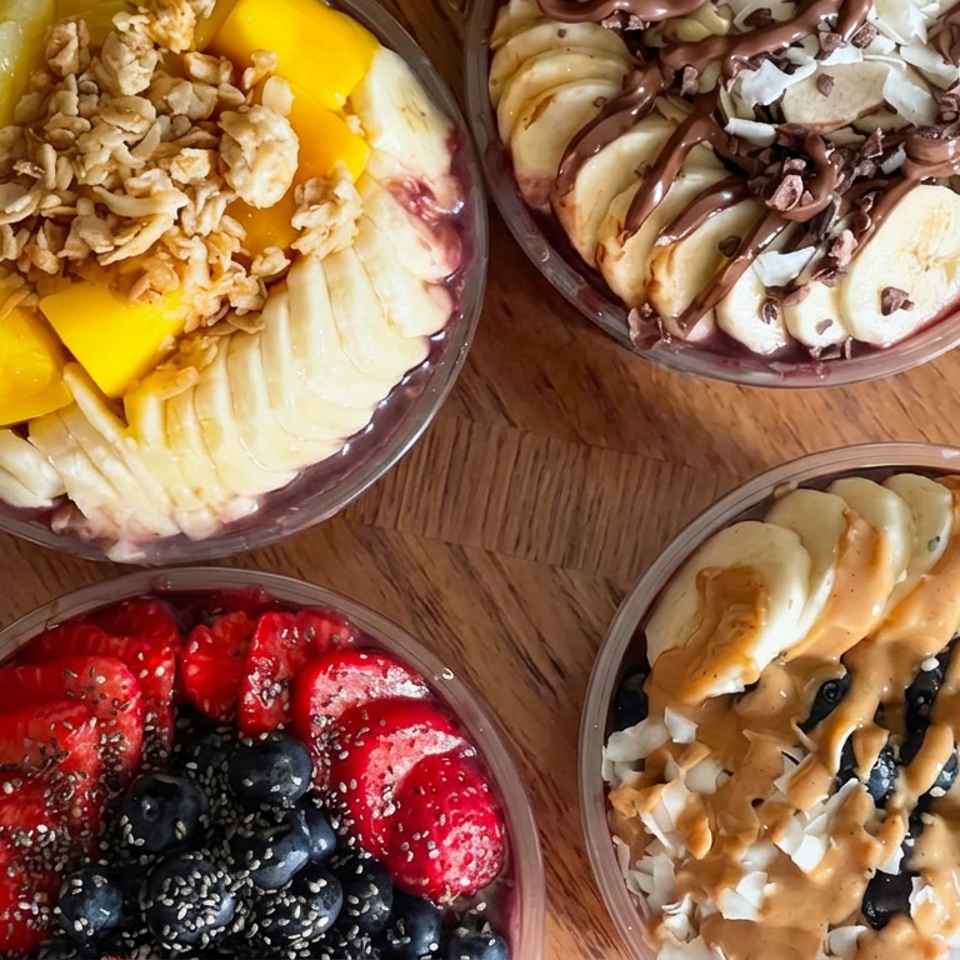 Four colorful smoothie bowls topped with sliced fruit, berries, granola, coconut, chia seeds, chocolate, and nut butter on a wooden table.